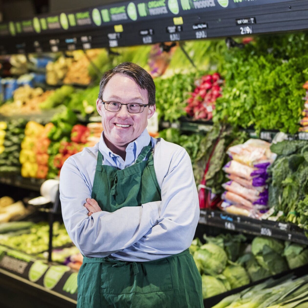 Man with down syndrome working in supermarket A mature man in his 40s with down syndrome working in a supermarket. He is wearing an apron, standing in the produce section of the store, looking toward the camera with his hands on his hips.